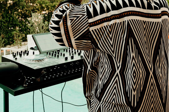 DJ performs at a poolside party during a warm summer evening with vibrant lights and music. DJ controls the equipment with one hand while the other rests on a mixer. Guests enjoy lively atmosphere