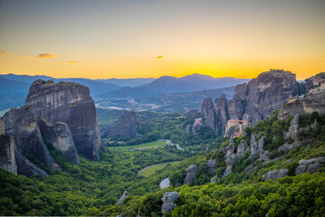Obraz premium Spectacular sunset view from a viewpoint overlooking Meteora in Greece. Warm golden and orange light bathes the towering sandstone rock formations and historic Orthodox monasteries