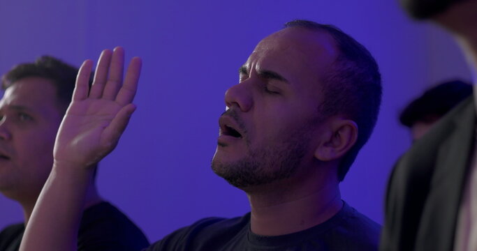 Hispanic man with raised hand and intense expression worshipping during evangelical Christian service, surrounded by fellow believers in prayerful focus