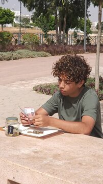 Teenage boy counting Euro banknotes and planning budget outdoors, financial literacy for youth concept. Focused curly-haired teen sitting at a table in a park, counting cash money next to a savings ja
