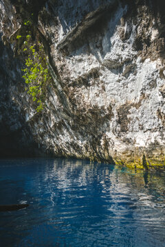 Clear blue water reflects off a plant-covered rock wall, Melissani Lake