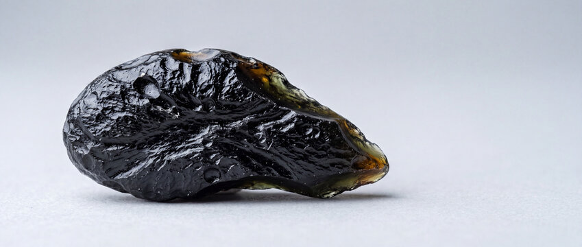 Raw black tektite glass stone specimen with shiny green edges on neutral background