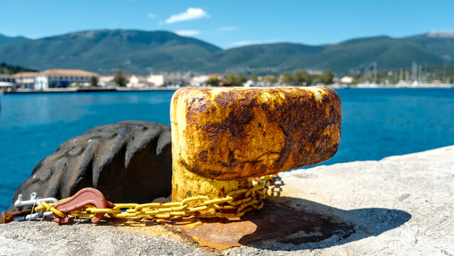 Rusty poller with yellow paint and chain on harbor wall, calm sea and mountains