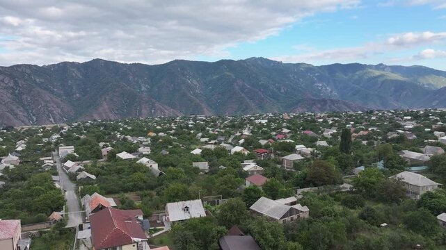 Beautiful aerial drone shot of the scenic Odzun village in Armenia, showing charming houses surrounded by lush green trees and majestic mountains under a bright summer blue sky.

