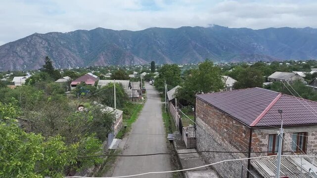 Beautiful aerial drone shot of the scenic Odzun village in Armenia, showing charming houses surrounded by lush green trees and majestic mountains under a bright summer blue sky.


