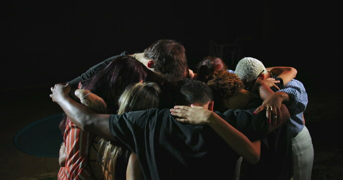Diverse group of people including Latin Hispanic and African American individuals standing in circle with arms around each other in emotional nighttime prayer gathering