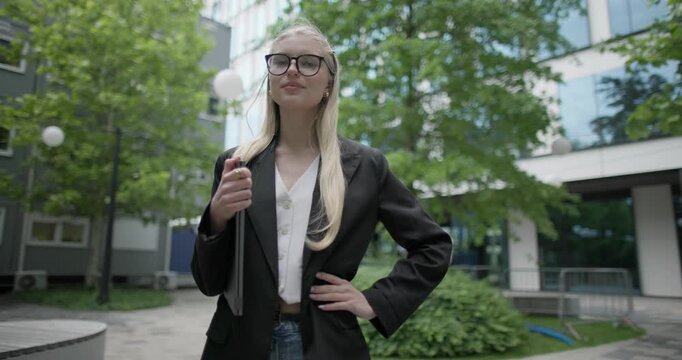 Young Caucasian female student wearing glasses in a modern urban environment among houses and trees standing in a park holding a laptop