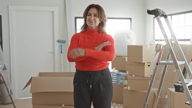 Woman with cardboard boxes smiling and arms crossed, standing among packed boxes, ladder and tape in building; confidence.
