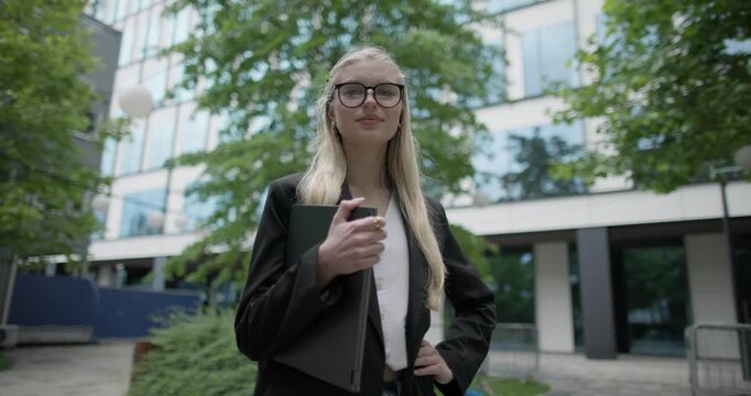 Young Caucasian female student wearing glasses in a modern urban environment among houses and trees standing in a park holding a laptop