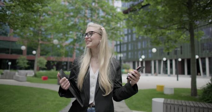 Young Caucasian female student wearing glasses in a modern urban environment among houses and trees walking in the park with a laptop and talking on the phone
