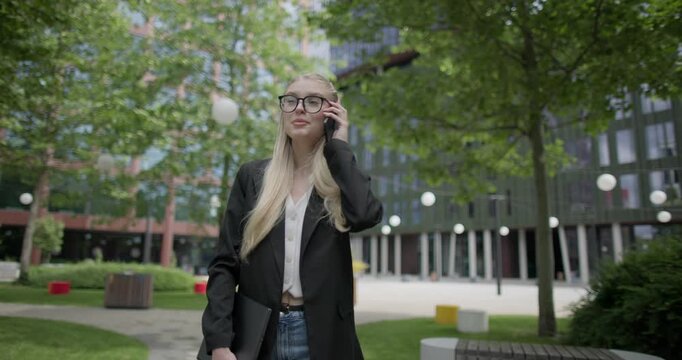 Young Caucasian female student wearing glasses in a modern urban environment among houses and trees walking in the park with a laptop and talking on the phone