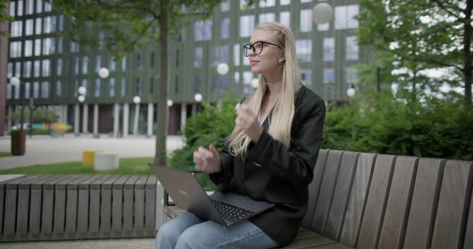 A young Caucasian female student wearing glasses in a modern urban environment among houses and trees sits on a bench in a park and works on a laptop