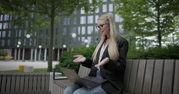 A young Caucasian female student wearing glasses in a modern urban environment among houses and trees sits on a bench in a park and works on a laptop conducting a video call, webinar or conference