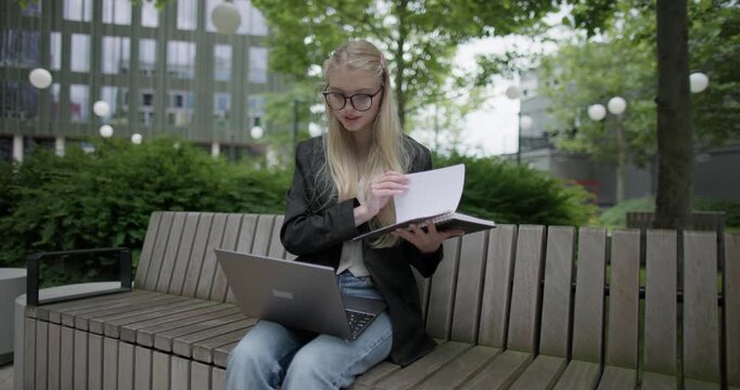 A young Caucasian female student wearing glasses in a modern urban environment among houses and trees sits on a bench in a park and works on a laptop reading notes from a notebook