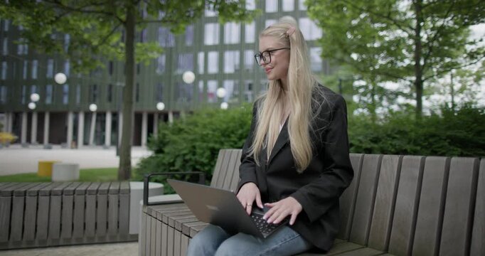 A young Caucasian female student wearing glasses in a modern urban environment among houses and trees sits on a bench in a park and works on a laptop