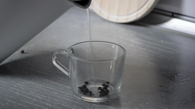 Close-up video of hot water being poured into a glass cup with tea leaves, as the leaves slowly unfurl and steep, creating a calming tea preparation scene.