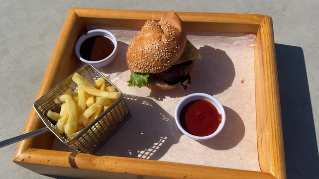 Lunch at snack bar: lunchbox with cheeseburger, fries, ketchup in sunlight by cafe window, casual outdoor dining.