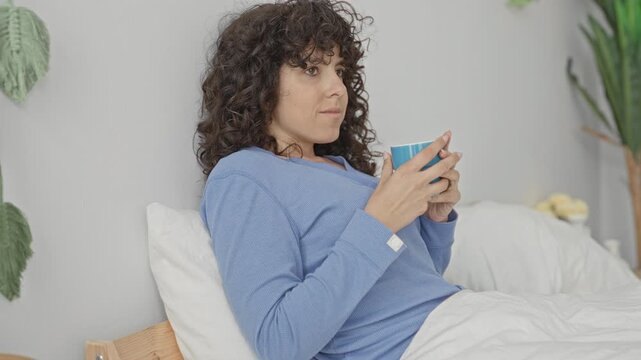 Young woman with curly hair holds blue coffee mug with bare hands while reclining on a bed in a studio; morning routine serenity.