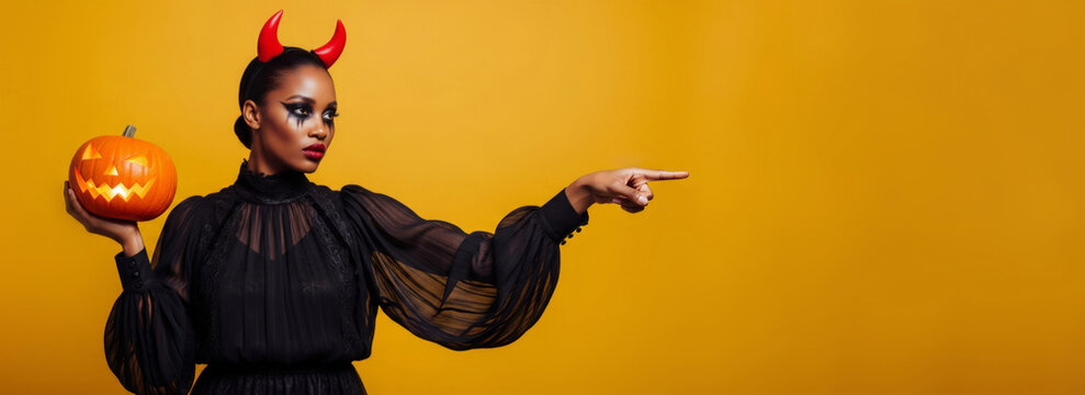 African American woman holding a jack-o-lantern and pointing, on a yellow background, dressed in a devil costume for a Halloween celebration. Banner template for holiday sale with copy space.