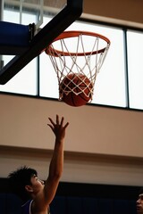 Fototapeta premium Player attempts basket in basketball match. Ball goes through net after successful shot. Athlete aims for score during game competition on indoor court.