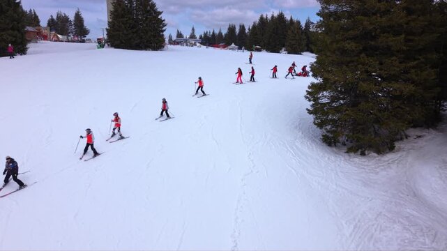 Aerial footage of a ski school lesson with beginner skiers practicing snowplow turns on a gentle slope at a mountain ski resort. The instructor leads the group downhill while other visitors enjoy wint