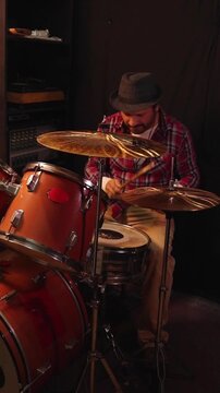 Medium shot of a male drummer in a plaid shirt and hat playing drums in a dark studio, vertical video