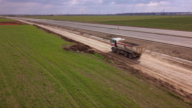 Aerial drone tracking shot of heavy dump truck transporting construction materials along temporary service road beside large highway expansion project with farmland, earthworks and infrastructure deve