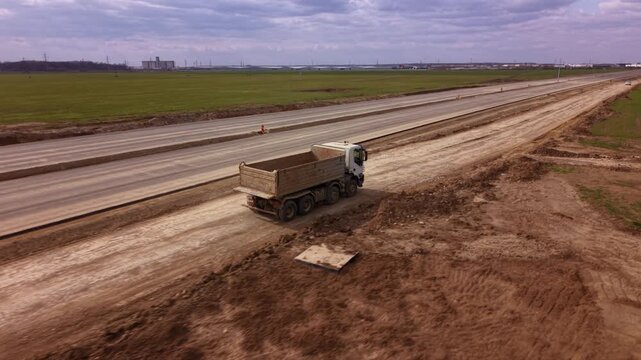Aerial drone tracking shot of heavy dump truck transporting construction materials along temporary service road beside large highway expansion project with farmland, earthworks and infrastructure deve