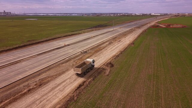 Aerial drone tracking shot of heavy dump truck transporting construction materials along temporary service road beside large highway expansion project with farmland, earthworks and infrastructure deve