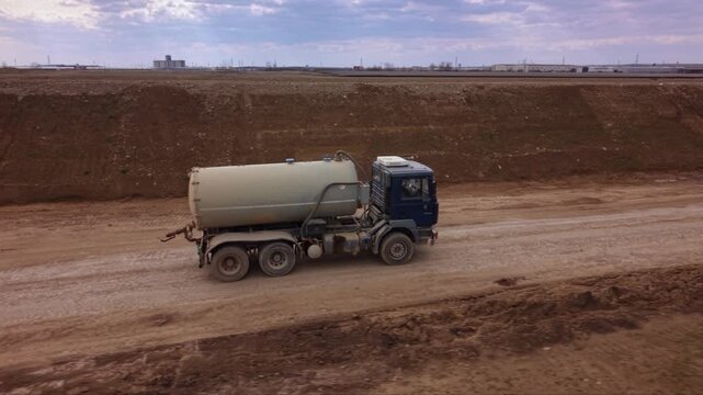 Aerial drone view of water tanker truck driving along unpaved road at large construction site, industrial vehicle supporting earthworks, road building, dust suppression and infrastructure development