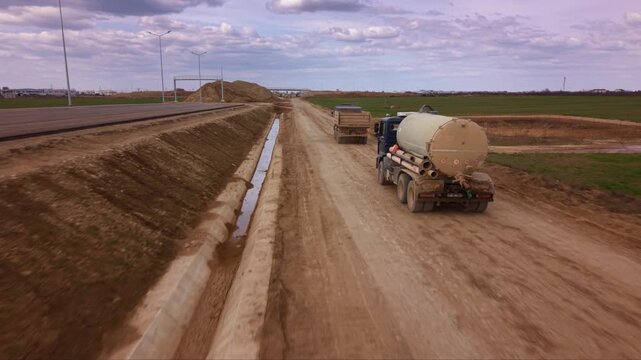 Aerial drone view of water tanker truck driving along unpaved road at large construction site, industrial vehicle supporting earthworks, road building, dust suppression and infrastructure development