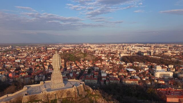 Aerial panoramic view of Alyosha Monument overlooking Plovdiv city at golden hour with sprawling urban skyline, Rhodope Mountains in the distance, historic landmark and dramatic evening clouds, Bulgar