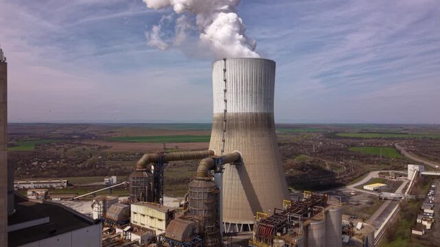 Close aerial drone view of a massive cooling tower releasing dense steam at a thermal power plant. Dramatic clouds and industrial landscape highlight electricity generation, energy infrastructure, and