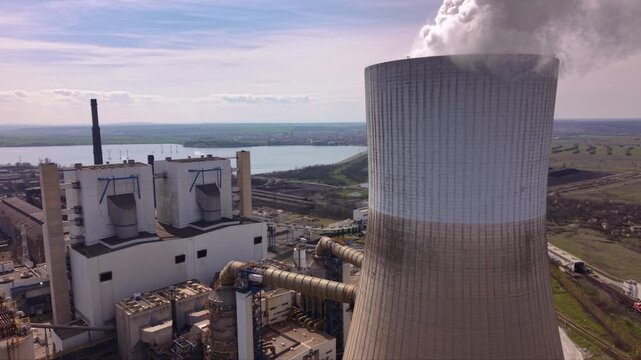 Close aerial footage of a massive cooling tower emitting dense white steam at an operating thermal power plant. The towering industrial structure dominates the landscape while showcasing electricity g