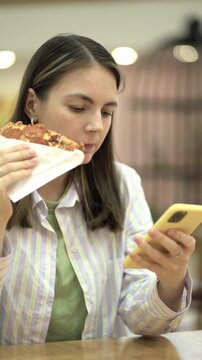 Young woman takes a bite of pizza wrapped in paper while scrolling on her yellow smartphone indoors. Casual multitasking with food and technology.