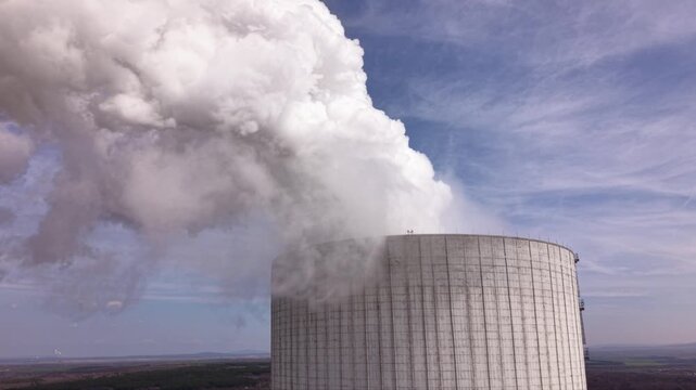 Close aerial footage of a massive cooling tower emitting dense white steam at an operating thermal power plant. The towering industrial structure dominates the landscape while showcasing electricity g