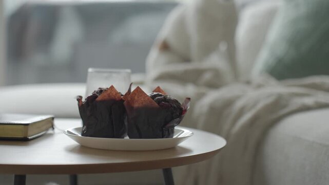 Hand sets plate of muffins beside glass of coffee on small table near sofa. Concept of coffee break and sweet snack