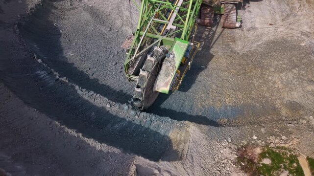 Aerial footage of a giant bucket wheel excavator continuously digging and loading rock in an active open pit quarry. The enormous mining machine transfers excavated material onto conveyor belts, showc