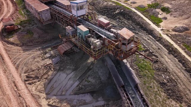 Aerial view of a long overland conveyor system transporting bulk material across a vast open-pit mining or industrial site, with transfer machinery, service roads and heavily disturbed terrain.