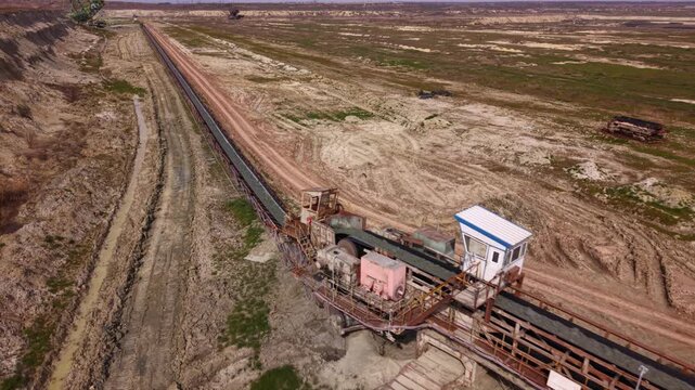 Aerial view of a long overland conveyor system transporting bulk material across a vast open-pit mining or industrial site, with transfer machinery, service roads and heavily disturbed terrain.