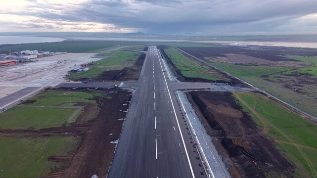 Aerial drone view of a newly constructed airport runway during expansion works under dramatic storm clouds. Fresh asphalt, taxiway intersections, construction activity, and surrounding airfield infras