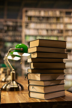 Close up of stacked books on desk in quiet university library with warm lighting and green lamp, educational environment with bookshelves and study materials, empty library study space.