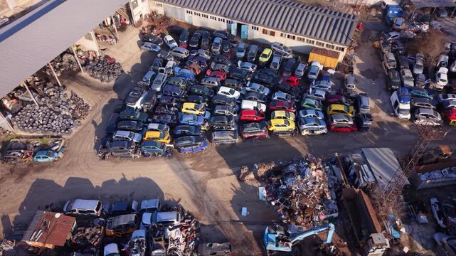 High-resolution aerial drone footage of an automotive scrapyard featuring hundreds of abandoned vehicles, scrap metal piles, industrial warehouses, and a vehicle recycling facility. Top-down view high
