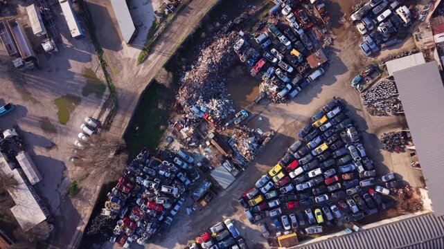 Top-down aerial view of a large vehicle scrapyard packed with rows of dismantled cars, scrap metal and automotive parts beside industrial buildings, showing vehicle recycling and waste processing.