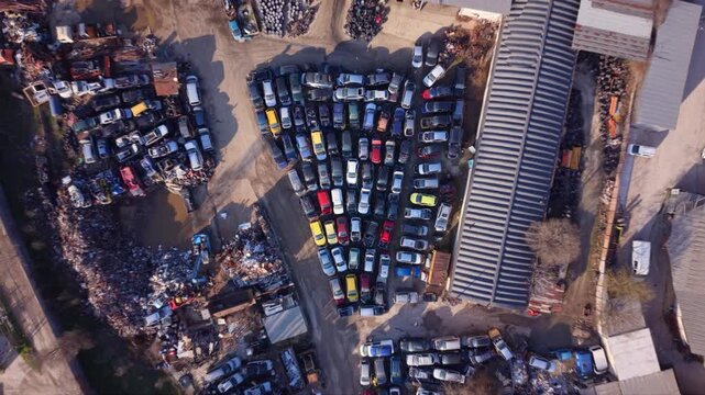 High-resolution aerial drone footage of an automotive scrapyard featuring hundreds of abandoned vehicles, scrap metal piles, industrial warehouses, and a vehicle recycling facility. Top-down view high