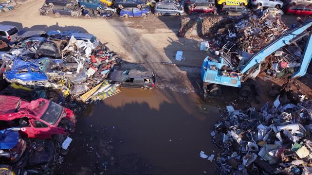 Top-down aerial view of a large vehicle scrapyard packed with rows of dismantled cars, scrap metal and automotive parts beside industrial buildings, showing vehicle recycling and waste processing.