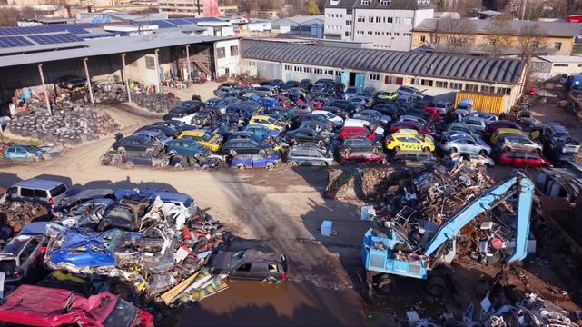 High-resolution aerial drone footage of an automotive scrapyard featuring hundreds of abandoned vehicles, scrap metal piles, industrial warehouses, and a vehicle recycling facility. Top-down view high