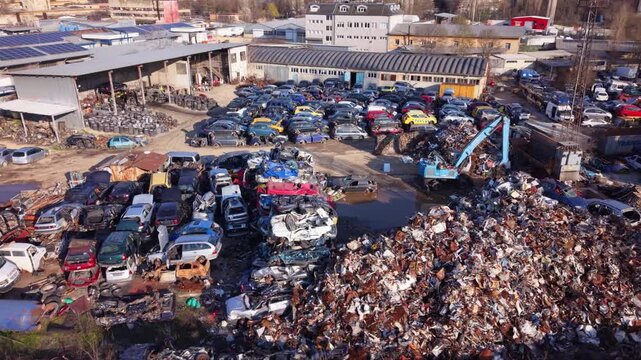 High-resolution aerial drone footage of an automotive scrapyard featuring hundreds of abandoned vehicles, scrap metal piles, industrial warehouses, and a vehicle recycling facility. Top-down view high