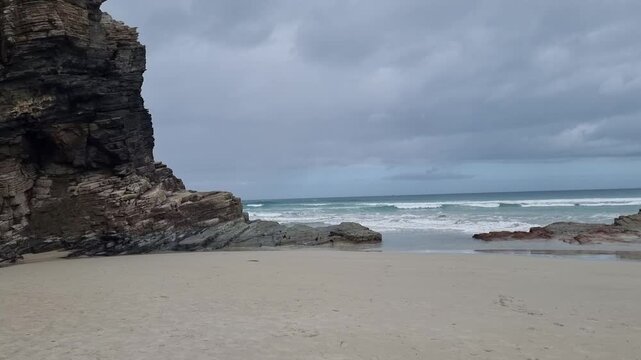Dramatic landscape of the natural sea arch at As Catedrais beach, Galicia, showcasing monumental, layered slate cliffs under an overcast sky.