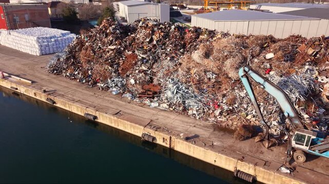 Aerial footage of a large scrap metal recycling facility at an industrial port, featuring massive stockpiles of ferrous and non-ferrous metal ready for processing. Warehouses, cranes and waterfront in